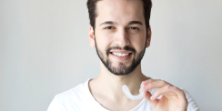 Junger Mann mit Bart lächelt und hält eine transparente Zahnschiene in der Hand vor hellem Hintergrund.