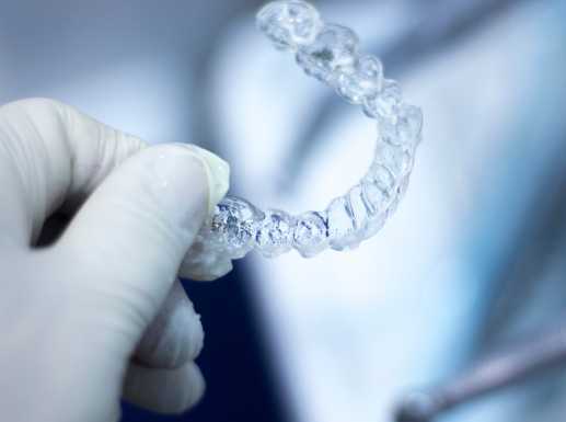 Eine Hand mit medizinischem Handschuh hält eine transparente Zahnschiene vor unscharfem Hintergrund.